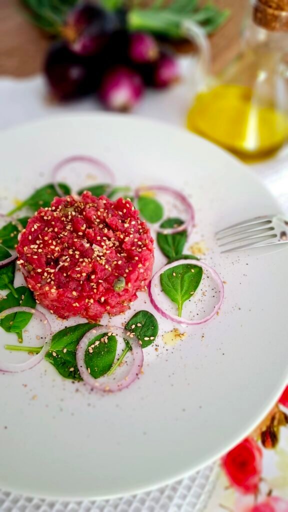 Tartare di chianina e scottona su piatto bianco a contrasto condita con capperi scalogno limone e olio e semini di sesamo finali con guarnizioni di spinacini e cipollotti a rondelle
