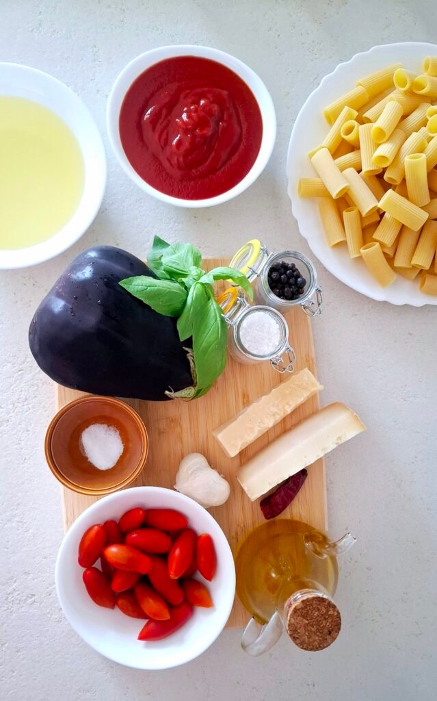 Imgredienti sul tagliere: pasta (i rigatoni o mezze maniche), la melanzana, la passata di pomodoro, i pomodorini, l'olio, l'olio di semi per friggere, il basilico, l'aglio, il pepe, il peperoncino, il sale grosso, il sale fino, il pecorino (o la ricotta salata) e il parmigiano.