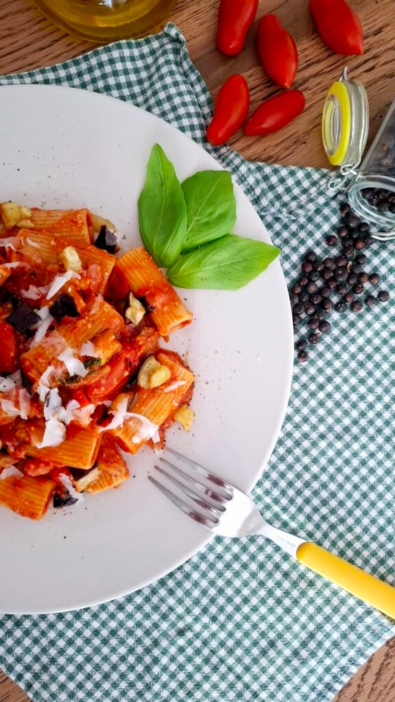 Rigatoni alla norma cremosissimi con melanzane basilico pepe nero peperoncino e pecorino su piatto chiaro tovaglietta quadri verdi e forchetta gialla