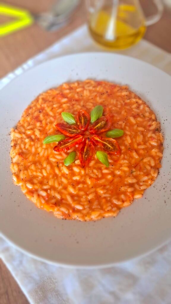 Risotto ai pomodorini confit cremoso con fiore di pomodorini e basilico al centro.Sfondo di posate verde acido e oliera.