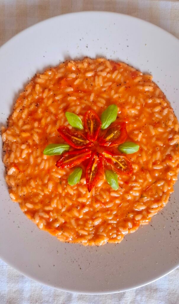 Risotto con pomodorini confit cremoso e filante con basilico e caciotta su piatto tondo r tovaglietta a quadri. Guarnito con un fiore di pomodorini e basilico.