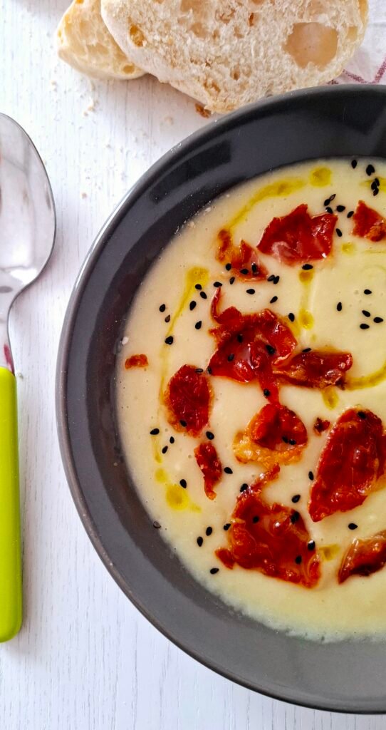 Primo piano su fondina grigia con vellutata di porri e cannellini cremosa, profumata con chips prosciutto crudo e sesamo nero. A fianco cucchiaio verde e sopra pane a fette