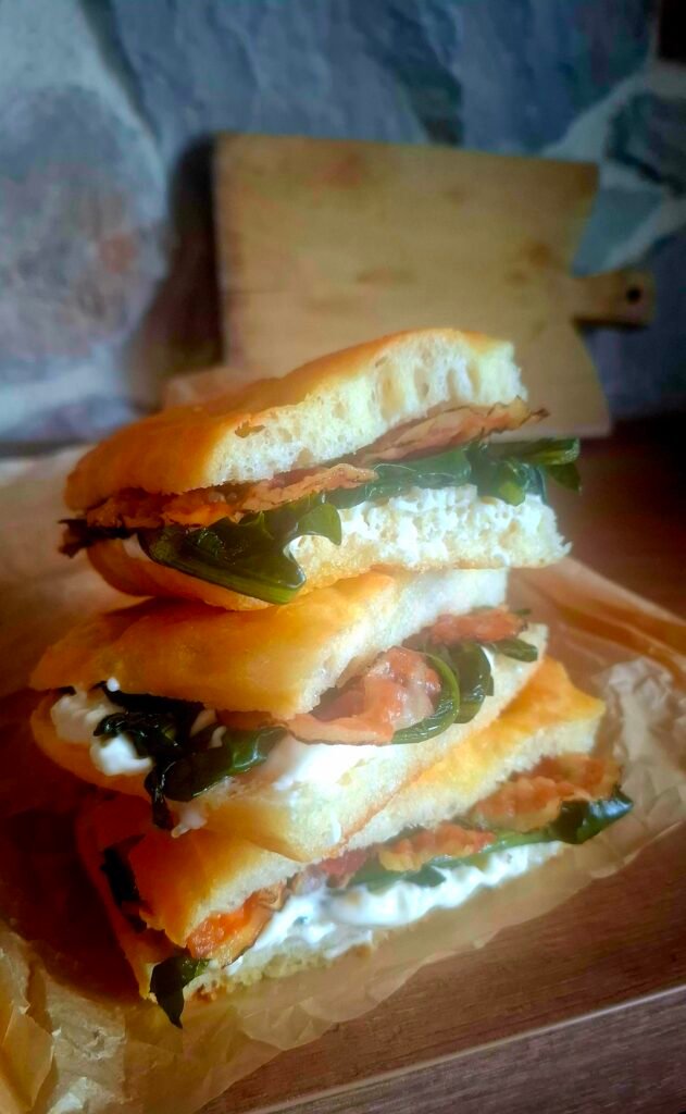 Focaccia morbida e alveolata con gli alveoli ben visibili messa una sopra l'altra con ripieno di stracchino, cicoria ripassata e pancetta pepata e miele. Sfondo rustico con legno, tagliere e pietra a vista.