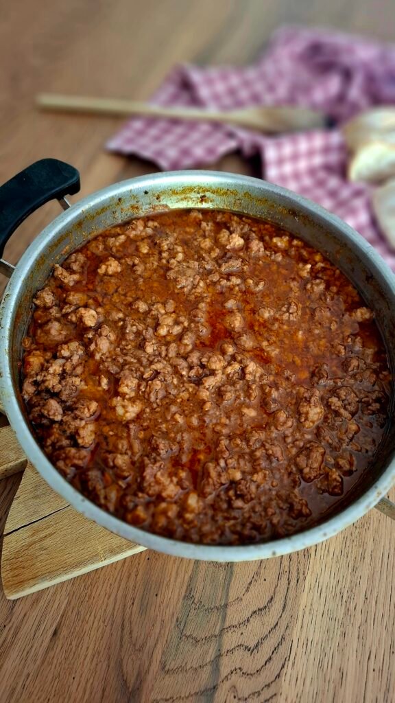 Ragù ricco e saporito in padella con vicino tovagliolo a quadretti rosso, cucchiaio in legno e pane a fette