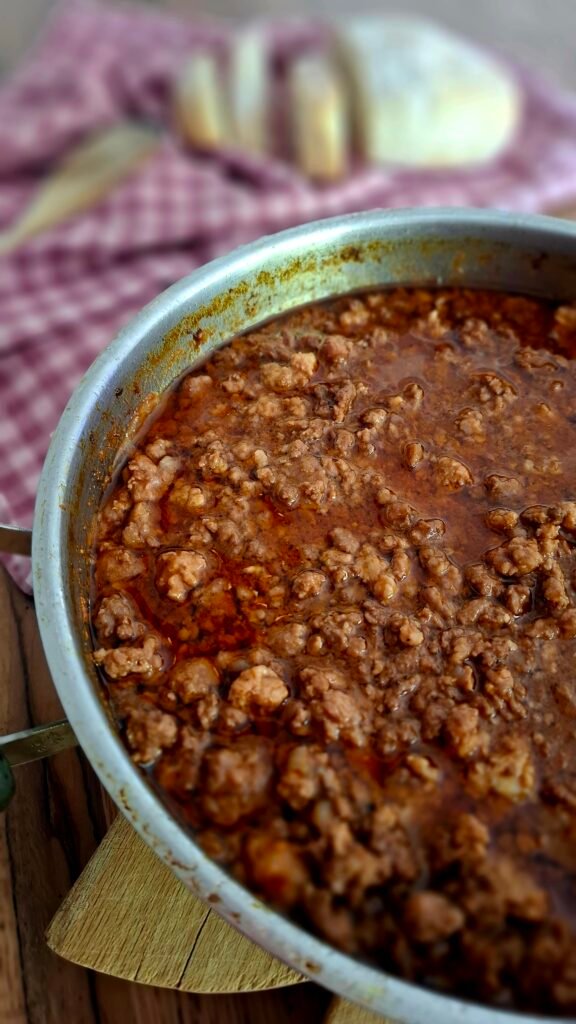 Ragù succulento, cremoso e corposo in padella su tagliere con sfondo di tovagliolo quadretti rossi, cucchiaio in legno e pane a fette.