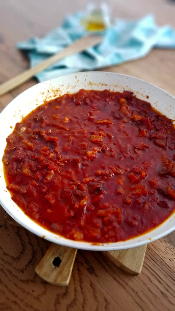 Ragù di prosciutto crudo morbido con passata di pomodoro, scalogno e vino bianco in padella su tagliere in legno; sfondo di tovagliolo a quadri verdi, cucchiaio in legno e oliera