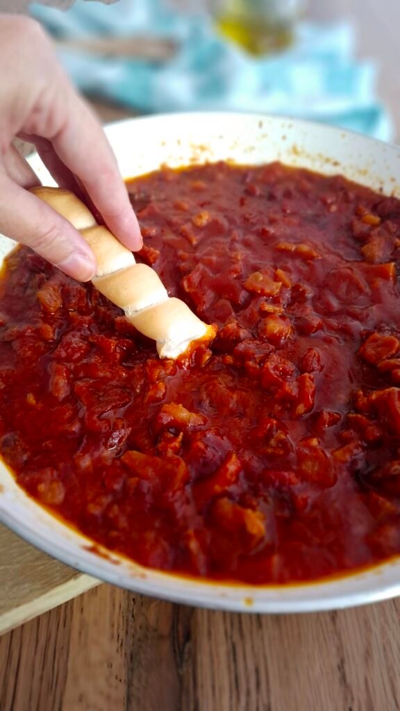 Crostino di coppia ferrarese intinto nel ragù di prosciutto crudo bolognese direttamente in padella. Sfondo di tovagliolo verde a quadro, cucchiaio in legno e oliera