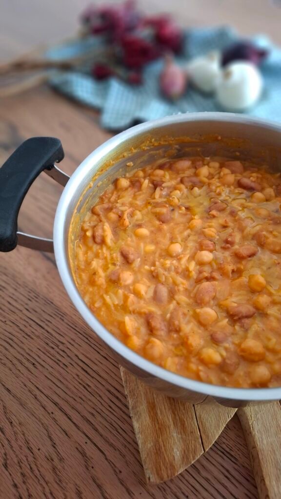 Padella con pasta e fagioli cremosa con ceci e cicerchie su tavolo in legno con sfondo di cipolle e peperoncini.