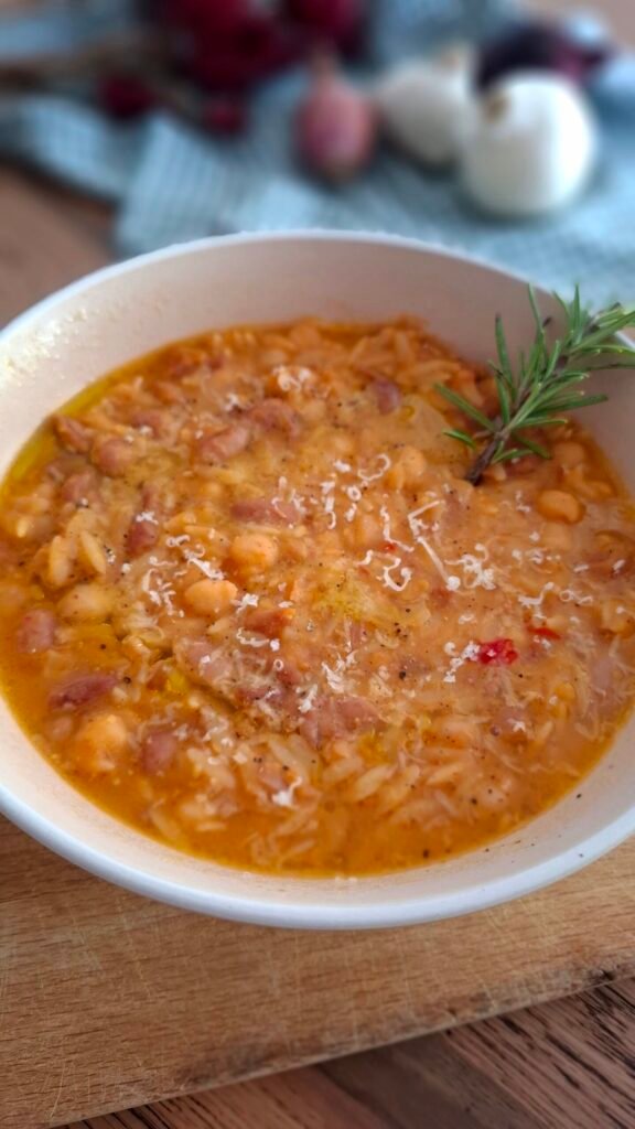 Pasta e fagioli con ceci e cicerchie presentata in fondina beige su tavolo in legno con sfondo di cipolle e peperoncini. Guarnita con olio, pepe, parmigiano e rametto di rosmarino.