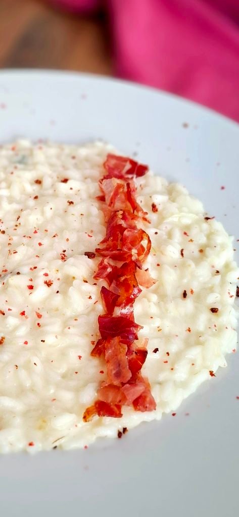 Risotto ai 4 formaggi (gorgonzola, taleggio, parmigiano, caciotta) cremoso e filante, con striscia di speck croccante e pepe rosa macinato. Sullo sfondo tovagliolo rosa dello stesso colore del pepe.