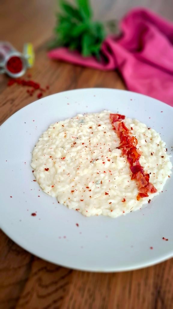 Piatto beige con risotto ai 4 formaggi all'onda. Guarnito con Speck croccante e pepe rosa.
Sullo sfondo tovagliolo rosa, mazzetto di erbe fresche e vasetto di pepe rosa