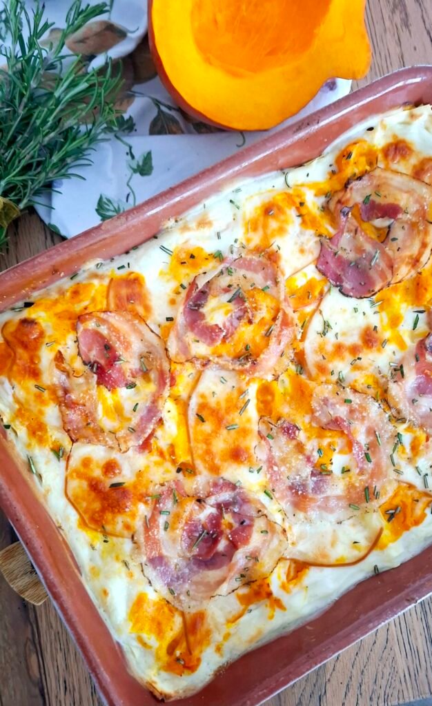 Lasagne autunnali sfiziose e cremose con zucca pancetta scamorza e rosmarino, in teglia su tavolo in legno con sfondo di rametto di rosmarino con fiocchetto arancio e zucche.
